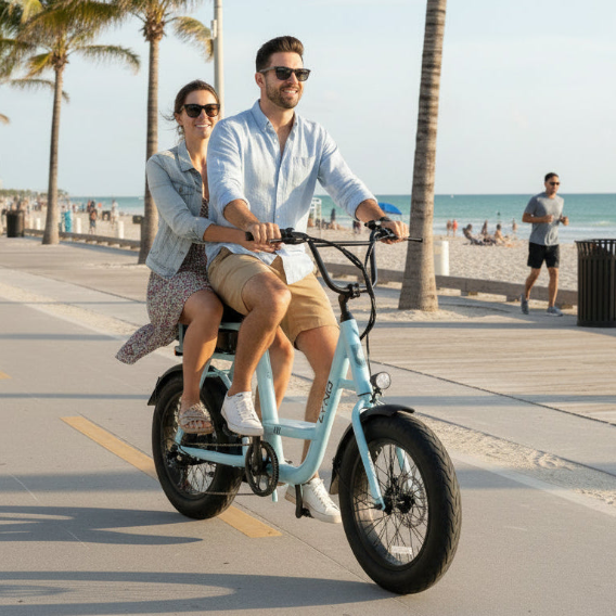 Man and woman riding a light blue electric bike on a boardwalk with palm trees and ocean in the background.